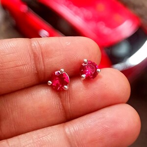 May include: A pair of stud earrings featuring round, vibrant red gemstones set in silver-colored metal. Each earring has three small prongs securing the gemstone. The earrings are displayed against a blurred background.