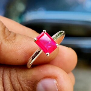 May include: A silver ring featuring a vibrant, rectangular, emerald-cut pink gemstone set in a four-prong setting. The band is smooth and polished, showcasing a clean, metallic finish that complements the bright, saturated color of the stone.