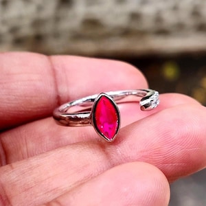 May include: A silver-toned open ring featuring a marquise-cut vibrant pink gemstone as the central focal point. The band is polished and includes a small cluster of clear, colorless stones set along one side of the open shank design.