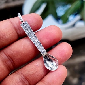 Op de afbeelding: Een zilveren lepelhanger met een kleine lus aan de bovenkant. De steel is versierd met twee rijen kleine, heldere edelstenen. De kom van de lepel is gepolijst en reflecterend.