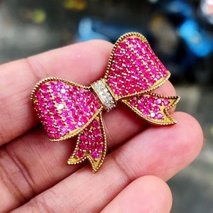 May include: A gold-coloured bow brooch, adorned with many small, bright pink gemstones. The centre of the bow features a section of clear, sparkling stones. The brooch has a detailed, textured edge.