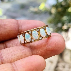 Puede incluir: Un anillo de oro con una serie de ópalos ovalados y pequeñas piedras preciosas transparentes. Los ópalos tienen una base blanca lechosa con destellos azules y verdes. El anillo se sostiene en una mano, mostrando el diseño detallado y la artesanía.