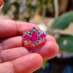 Könnte beinhalten: Ein silberfarbener Ring mit einem zentralen, leuchtend pinkfarbenen Edelstein im Marquise-Schliff, flankiert von zwei kleineren pinkfarbenen Steinen, umgeben von einer Ansammlung kleiner, klarer, funkelnder, rund geschliffener Steine in einer Metallfassung.