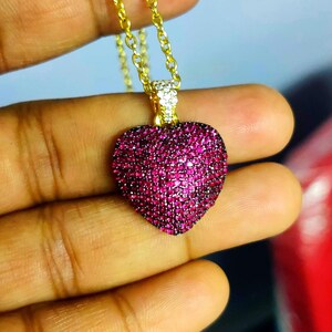May include: A heart-shaped pendant necklace featuring a vibrant fuchsia-pink pavé surface. The heart is suspended from a delicate gold-tone chain, with a small, clear crystal-encrusted bail connecting the pendant to the link chain structure.
