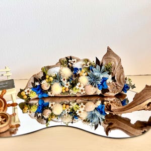 May include: A decorative arrangement featuring a large seashell filled with blue and white dried flowers, small white seashells, and greenery. Beside it are two wooden ship wheel ornaments and a small white chair with a sign reading ST-28.