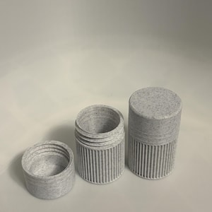 Puede incluir: Juego de tres piezas de recipientes cilíndricos grises moteados. El juego incluye una tapa, una base con estrías verticales y un recipiente más alto con una tapa moteada a juego. Los recipientes están hechos de un material gris claro con motas grises más oscuras.