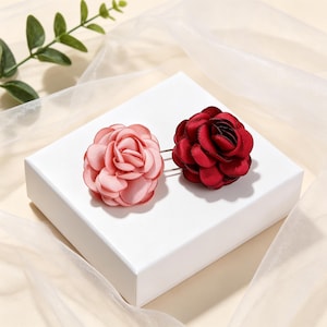 May include: Two fabric flower pins, one in a soft pastel pink and the other in a deep burgundy red. Each flower features layered, rounded petals arranged in a circular, blooming rose shape, attached to a thin silver-toned metal pin backing.