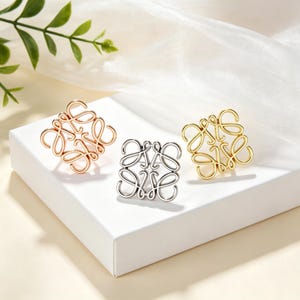 Puede incluir: Tres broches cuadrados con un diseño de logotipo cursivo intrincado e interconectado. Un broche es de oro rosa, otro es de tono plateado y el tercero es de tono dorado. Cada pieza está hecha de metal pulido con una estructura de alambre continuo.
