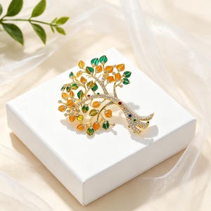Puede incluir: Un broche de árbol en tono dorado con ramas adornadas con pedrería transparente y hojas esmaltadas en tonos naranja, amarillo y verde. El tronco y las ramas principales están incrustados con pequeños cristales claros y piedras rojas y azules.