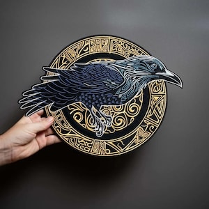 May include: A circular embroidered patch featuring a black raven with blue and white feather detailing, layered over a gold and black circular emblem with intricate geometric and swirling patterns. The design is stitched on a black fabric base.