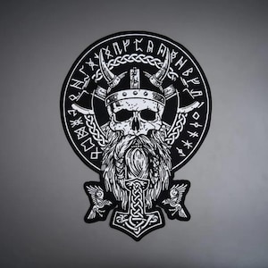 May include: A black and white circular patch featuring a detailed illustration of a bearded skull wearing a horned helmet. The design includes intricate knotwork, runic symbols around the border, and a central hammer emblem at the bottom.