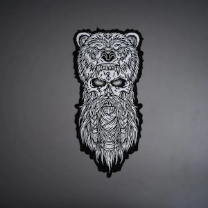May include: A black and white embroidered patch featuring a detailed illustration of a bearded warrior wearing a bear head as a hood. The design includes intricate line work on the bear fur, the skull-like face, and the braided beard.