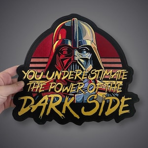 May include: A large embroidered patch featuring a stylized helmeted figure in red, blue, and black tones against a sunset background. The text "YOU UNDERESTIMATE THE POWER OF THE DARK SIDE" is stitched in bold, golden yellow lettering across the center.