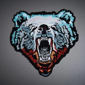 Puede incluir: Un parche bordado que muestra la cabeza de un oso rugiendo. El diseño utiliza hilos de color azul claro, blanco y rojo oscuro sobre un fondo negro. El oso tiene la boca abierta mostrando los dientes y un interior oscuro, con costuras detalladas.