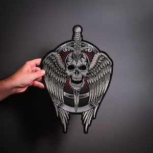 Könnte beinhalten: Ein großer gestickter Aufnäher mit einem zentralen menschlichen Totenkopf mit detaillierter Schattierung, der hinter einem vertikalen Schwert platziert ist. Das Design umfasst weit ausgebreitete gefiederte Flügel vor einem kreisförmigen Hintergrund mit roten Strahlen.