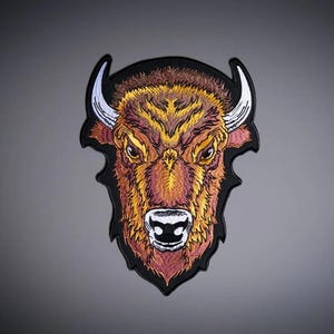 Op de afbeelding: Een geborduurde patch met de kop van een bizon in tinten bruin, oranje en geel. De bizon heeft witte hoorns met zwarte omlijningen en een zwarte neus. Het gehele ontwerp is omlijst door een dikke zwarte rand tegen een grijze achtergrond.