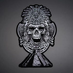 Puede incluir: Un parche bordado en blanco y negro que presenta una calavera detallada con un tocado ornamentado con motivos de calendario circulares. Debajo de la calavera hay una representación de una estructura de pirámide escalonada con hilos de alto contraste.