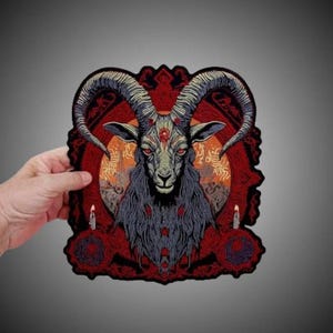 May include: A rectangular embroidered patch featuring a central goat head with large curved horns, red eyes, and a red circular symbol on its forehead. The design includes a dark red background with two lit candles and intricate ornamental patterns.