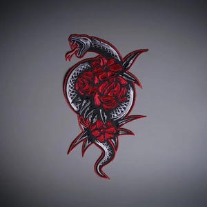 Pode incluir: Um emblema bordado apresentando uma cobra preta e branca enrolada em torno de três rosas vermelhas vibrantes. A cobra tem a boca aberta com uma língua vermelha, e todo o design é contornado por uma borda vermelha sobre um fundo cinza escuro.
