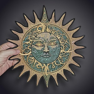 Puede incluir: Un parche bordado circular que presenta un sol estilizado con un rostro humano. El diseño incorpora intrincados patrones de remolinos en tonos verde azulado y verde, rodeados por rayos dorados radiantes con un borde negro.