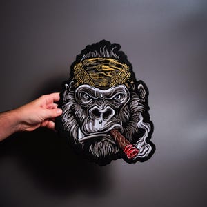 Könnte beinhalten: Ein bestickter Aufnäher mit einer detaillierten Illustration eines Gorillas, der ein goldenes Bandana mit Muster trägt. Der Gorilla hat einen ernsten Gesichtsausdruck und hält eine braune Zigarre im Mund, aus der weißer Rauch aufsteigt.