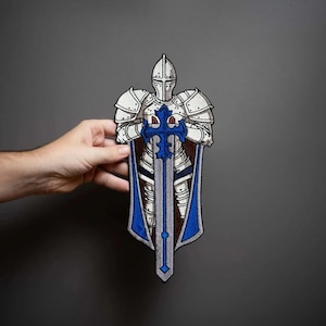 May include: An embroidered patch featuring a knight in silver plate armor standing with hands resting on a large, upright blue and silver sword. The knight wears a blue cape and is depicted in a detailed, upright stance with a closed helmet.