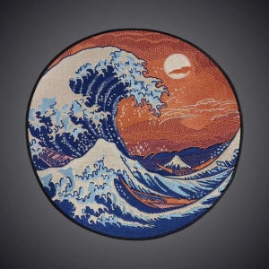 Puede incluir: Un parche bordado circular que presenta una ola oceánica estilizada en tonos azules y blancos, sobre un fondo cálido de color naranja y rojo con un sol pálido. El diseño incluye pequeñas embarcaciones navegando en el agua y una montaña.