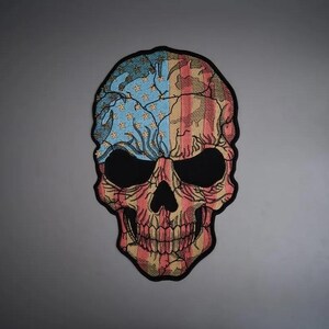 May include: A skull-shaped embroidered patch featuring a stylised American flag design. The left side displays blue with gold stars, while the right side features red and white vertical stripes. The entire design has a black border outline.