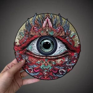 May include: A circular embroidered patch featuring a central realistic human eye with a green iris and black pupil. The surrounding area is filled with intricate, dense patterns in shades of red, teal, gold, and black, creating a complex design.