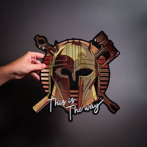 May include: An embroidered patch featuring a gold-colored helmet centered over a circular red and orange striped background. Crossed behind the helmet are a wrench and a hammer. The white text "This is The way" is stitched at the bottom.