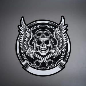 Puede incluir: Un parche circular en blanco y negro que presenta una calavera central con casco y gafas. Dos pistones de motor cruzados se sitúan bajo la calavera, flanqueados por grandes alas emplumadas. Un borde de cadena rodea el diseño con una cinta.