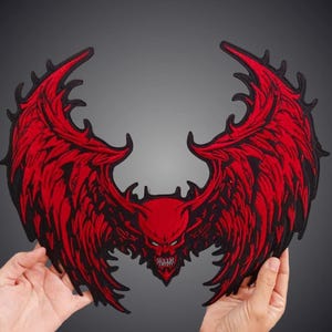 Puede incluir: Un parche bordado grande que presenta una cabeza de demonio roja central con dientes afilados y cuernos, flanqueada por dos alas rojas detalladas y expansivas con contornos negros. El diseño es simétrico y muestra costuras intrincadas.