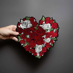 Puede incluir: Un parche bordado en forma de corazón que presenta un arreglo denso de rosas rojas y blancas con hojas verdes. El diseño se compone de múltiples flores de rosa de tamaños variados, todas delineadas con hilo negro sobre un fondo oscuro.