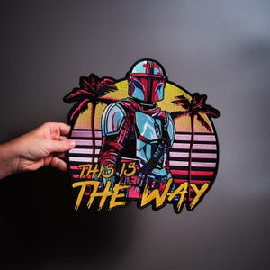 Op de afbeelding: Een geborduurde patch met een Mandalorian-personage in een lichtblauwe en rode helm en harnas. De achtergrond toont een zonsondergang met silhouetten van palmbomen en horizontale strepen in roze en geel. De tekst luidt THIS IS THE WAY in het geel.