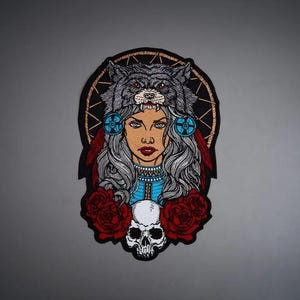 May include: A rectangular embroidered patch featuring a central illustration of a person wearing a wolf headdress, long gray hair, and blue clothing. The design includes a circular web background, two red roses, and a white skull at the bottom.