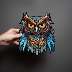 May include: An embroidered patch featuring a detailed owl design with vibrant colors including bright orange, deep blue, and gold. The owl has large, piercing yellow eyes, a sharp beak, and intricate feather patterns throughout its entire body.