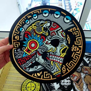 Puede incluir: Un parche bordado circular con un diseño de calavera estilizada en hilo rojo, amarillo, gris y azul. La calavera está rodeada por un borde negro con patrones geométricos dorados y un arco superior de motivos de ojos centrados en azul sobre fondo gris.