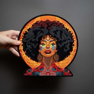 May include: A circular embroidered patch featuring a portrait of a person with a large afro hairstyle, wearing large circular earrings. The design includes vibrant orange, red, and teal colors with intricate patterns and a black border.