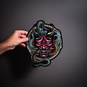 May include: A circular embroidered patch featuring a red Japanese Hannya mask with sharp horns and bared teeth, encircled by a green serpent and stylized blue water waves, all framed by a thick, textured rope border on a black background.