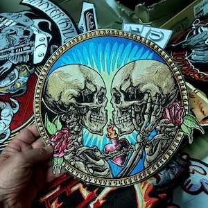 May include: A circular embroidered patch featuring two beige human skulls facing each other, surrounded by a bright blue sunburst background. The design includes red roses with green leaves and a central red heart with flames and thorns.