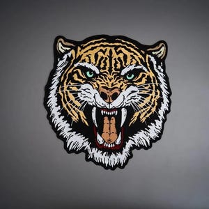 May include: A large embroidered patch featuring the head of a snarling tiger with its mouth open, revealing sharp teeth. The design uses vibrant orange, black, and white thread with striking green eyes, all outlined by a thick black border.