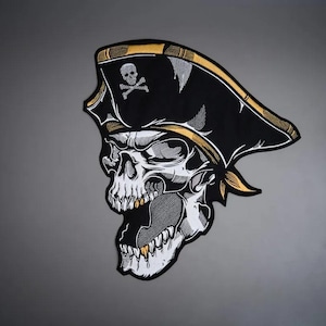 May include: An embroidered patch featuring a skull wearing a black pirate hat with a gold trim and a small skull and crossbones emblem. The skull has a wide-open jaw, visible teeth with gold accents, and is rendered in black, white, and gray tones.