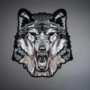 May include: An embroidered patch featuring the head of a snarling wolf. The design uses black, white, and grey thread to create detailed fur textures and facial features, including sharp teeth and an open mouth, finished with a black border.