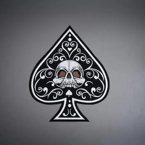 Op de afbeelding: Een zwart-wit schoppen speelkaartsymbool met een gedetailleerde menselijke schedel in het midden. Het ontwerp bevat ingewikkelde witte filigraan krulpatronen rondom de schedel en een klein schoppenicoon direct onder de kaaklijn.