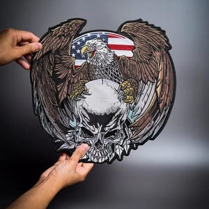 Puede incluir: Un gran parche bordado circular que presenta un águila calva detallada con alas marrones extendidas, agarrando un cráneo humano blanco con sus garras amarillas. Detrás del águila hay un gráfico circular de la bandera estadounidense.