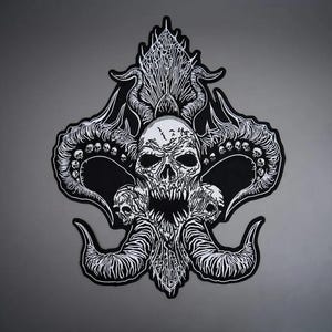 Könnte beinhalten: Ein schwarz-weiß bestickter Aufnäher mit einem zentralen Totenkopf mit spitzen Zähnen, flankiert von zwei kleineren Totenköpfen. Das Design zeigt große, geschwungene, texturierte Hörner und komplexe, flammenartige Muster, die nach oben und unten ragen.