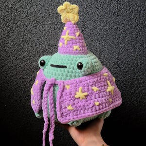 Puede incluir: Peluche de rana de ganchillo con sombrero y capa de mago. La rana es de color verde claro con ojos negros y boca negra. El sombrero y la capa son morados con estrellas amarillas. El sombrero tiene una flor amarilla encima.
