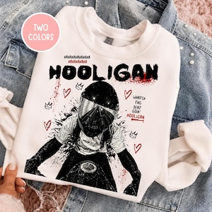 May include: White sweatshirt featuring a motorcycle graphic and the word "HOOLIGAN" in black and red. Additional text and heart and crown graphics are included. The sweatshirt is displayed on a denim jacket. The text "TWO COLORS" is in a pink circle.