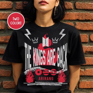 Op de afbeelding: Zwart T-shirt met een witte, verweerde opdruk met de tekst "THE KINGS ARE BACK" boven het woord "ARIRANG". Het ontwerp bevat rode vlammen, kronen en bliksemschichten. Een cirkelvormig label geeft twee kleuropties aan.