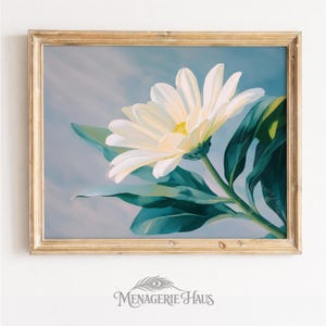 May include: A framed floral print depicting a large white daisy with a yellow centre and green leaves against a soft blue backdrop. The artwork is in a gold-coloured wooden frame. The Menagerie Haus logo is at the bottom.